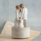 Faceless couple figurines sitting down holding hands in all white sitting on round white pedestal with music sheet and blue background