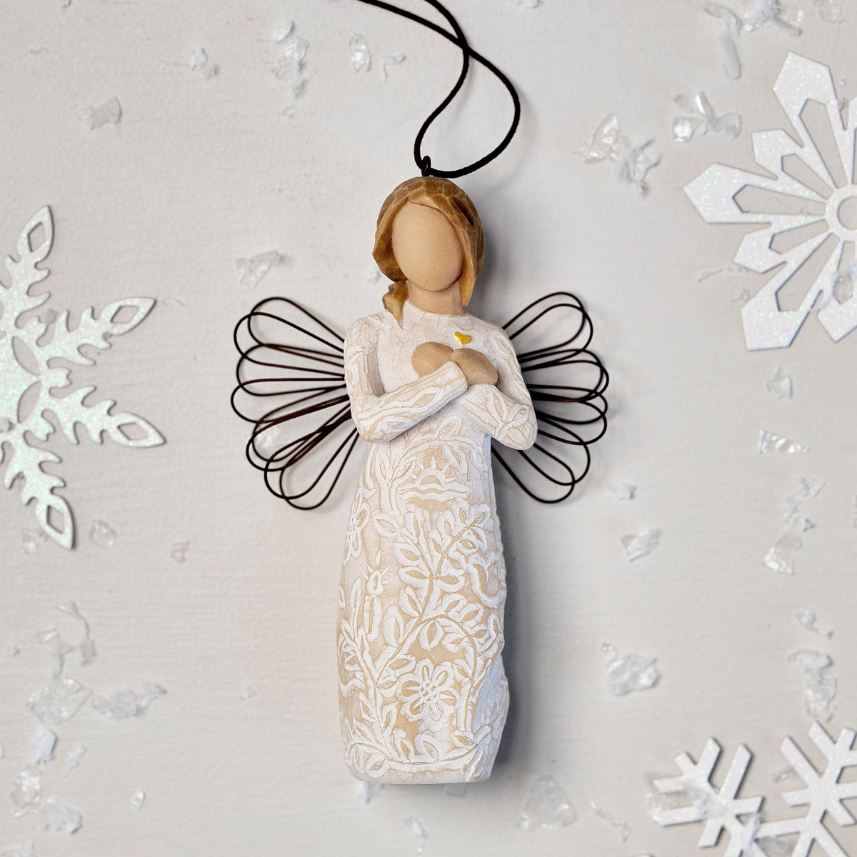 Willow Tree Remembrance Ornament, 4.5”h hand-painted resin angel figure in cream dress carved with symbols of nature, wire wings, crossed hands under small gold-leaf heart, brown elastic cord, laying amongst snowflakes