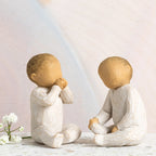 Willow Tree Two Together, two hand-painted seated resin figures of babies, tallest 2", one with hands at mouth, one with hands on legs, both in cream onsies, in front of pastel painting