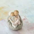 Willow Tree My new baby (blush), 2.5”h hand-painted resin figure of toddler in blush pink onesie holding infant in cream, cuddled together in a beanbag, in front of pastel painting