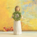Willow Tree Magnolia, 5.5”h hand-painted standing resin figure in cream dress, holding green magnolia leaf wreath, in front of dark yellow painting