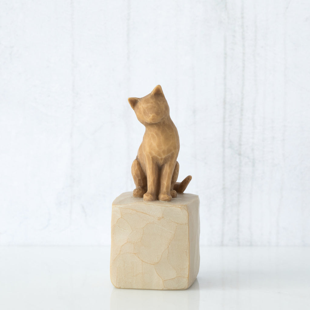 Figure of orange cat with tilted head perched atop a cream colored rectangular pedestal, facing front