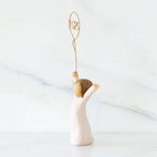 Facing left: standing figure in cream dress, arms raised, holding gold wire balloon with 2025 written in gold wire inside