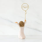 Back view: standing figure in cream dress, arms raised, holding gold wire balloon with 2025 written in gold wire inside