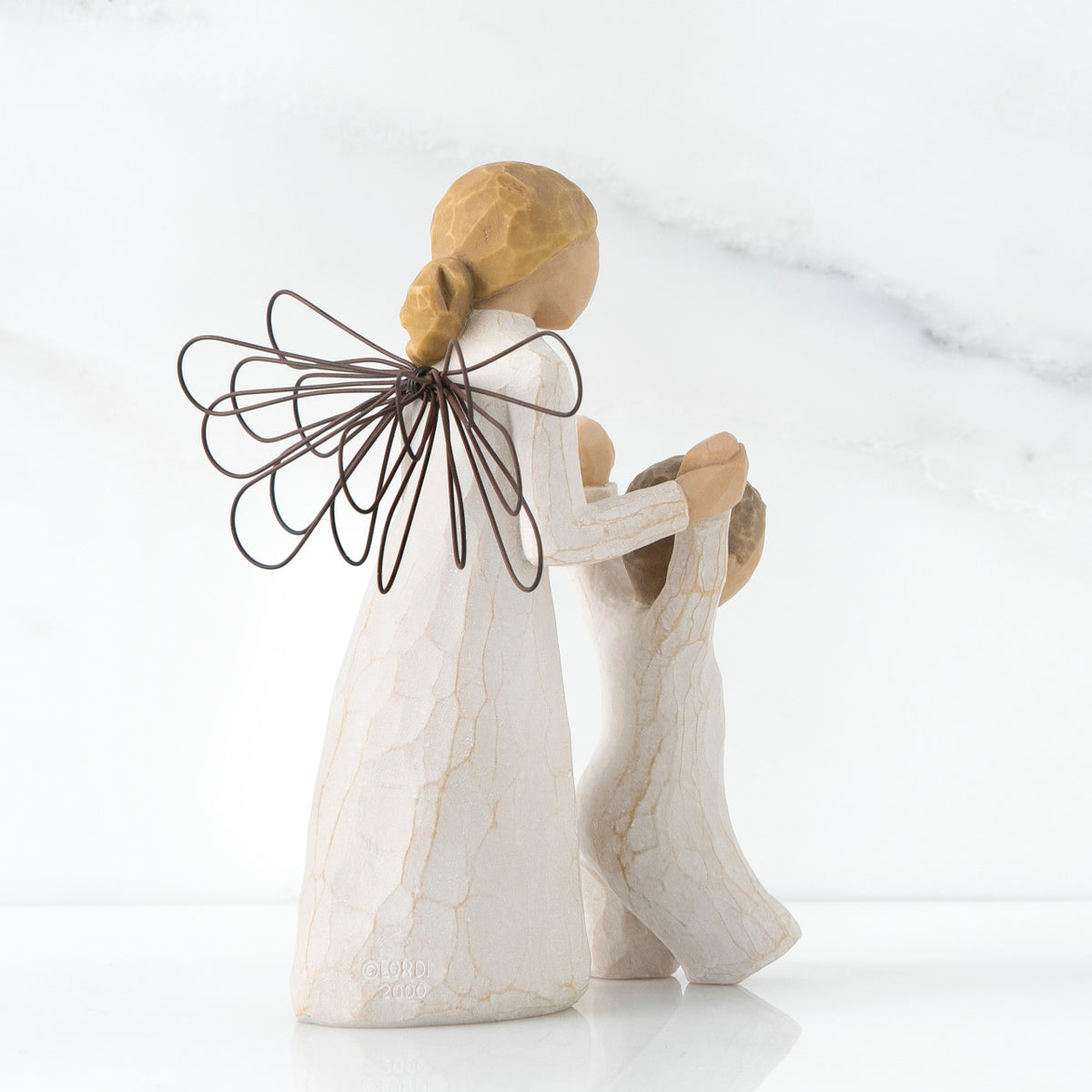 Blonde angel figurine in white dress, black wire wings, facing the back holding baby figurine's hands up