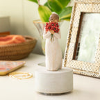 Willow Tree Surrounded by Love Musical, 7.5”h hand-painted resin figure in cream dress holding large bouquet of red-orange poppies up to face, atop round, cream base containing musical element, on desk with photos