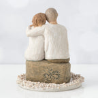 Back view of man and woman figurine in white with arms around another sitting on rock under round white flower plaque