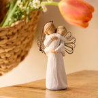 Willow Tree Angel's Embrace Ornament, 5"h hand-painted resin figure in cream dress with wire wings, holding baby in arms, brown elastic cord, standing on shelf with tulips