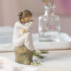 Willow Tree Guardian, 6”h hand-painted resin figure in cream dress, seated on gray rock, holding newborn baby in cream blanket, on table with glass vase