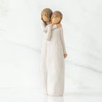Two faceless woman figurines standing wearing white dresses hugging one another