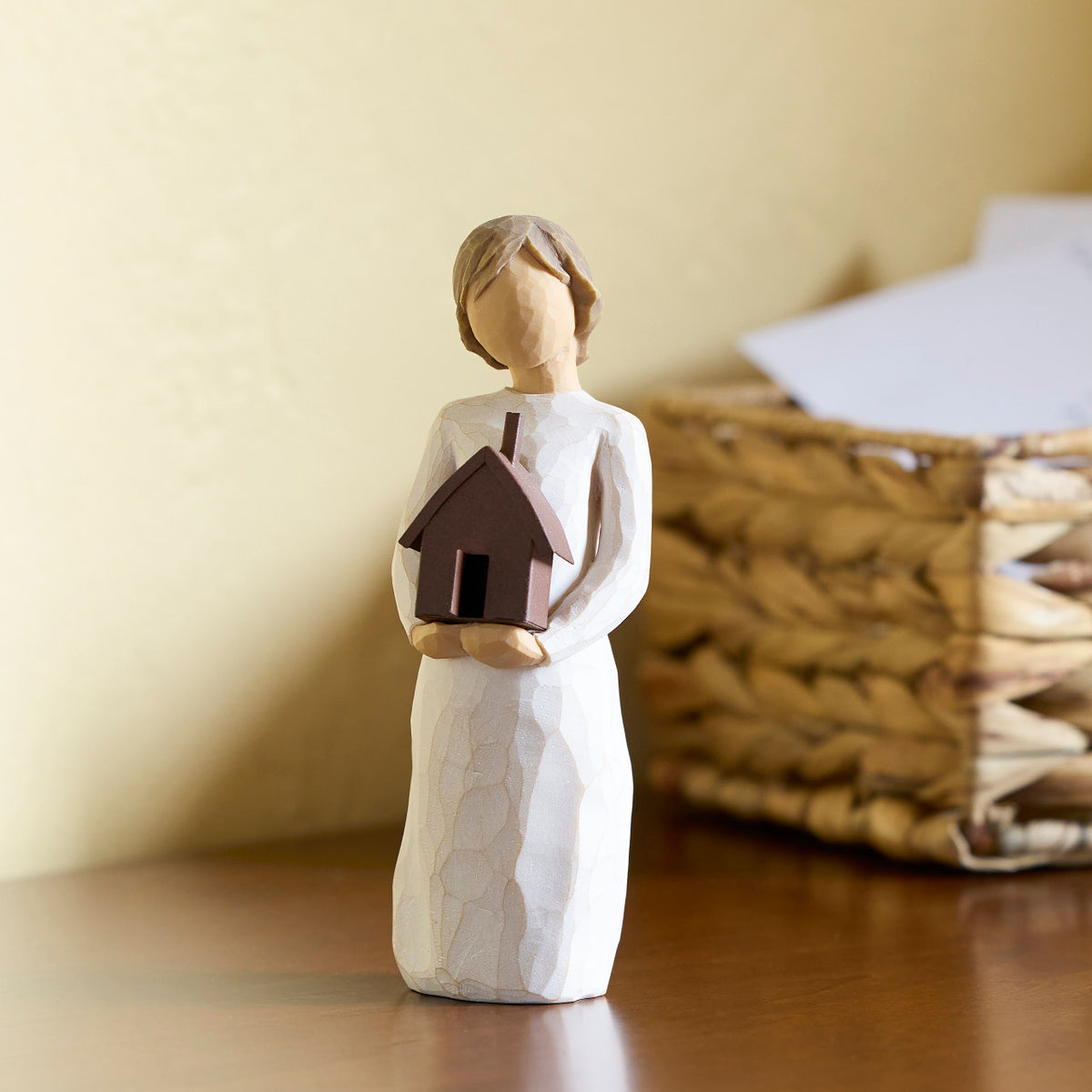 Willow Tree Mi Casa, 5.5”h hand-painted standing resin figure in cream dress, holding dimensional metal house in her arms, on table with basket