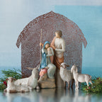 Nativity scene figurines sitting down holding baby next to lambs with brown barn in the background