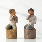 Two options (lighter skin and darker skin) of boy figurine kneeling on rock, holding wire circle with ‘love you’ inside