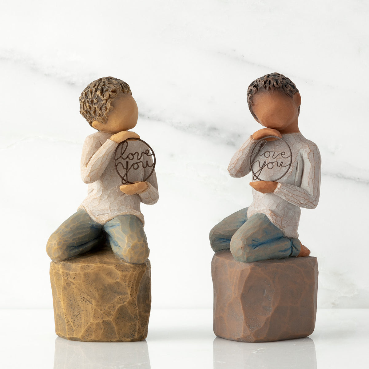 Two options (lighter skin and darker skin) of boy figurine kneeling on rock, holding wire circle with ‘love you’ inside
