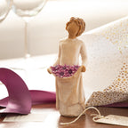 Willow Tree Simple Joys, 5.5”h hand-painted standing figure in cream dress, holding up skirt filled with purple daisies, on table with purple ribbon