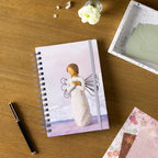 Willow Tree Remembrance Notebook, 6.5"w x 8.5"h, hard-cover, spiral-bound, 240 lined pages, front cover features Remembrance figure on background of purple-blue painting, closed on desk