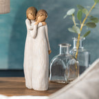 Willow Tree Chrysalis, 9”h hand-painted standing resin figure of two women in cream dresses; one standing behind and embracing the other, on table with glass vases