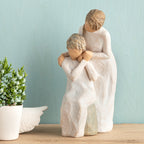 Willow Tree Loving My Mother, 6.5”h hand-painted resin figure of seated older woman with younger woman standing at her shoulder, both in cream dresses, hands clasped, in front of muted teal painting