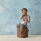 Willow Tree Love you too (darker skin), 4.5”h hand-painted resin figure of young child in cream shirt and jeans, kneeling on rock, holding wire circle with ‘love you’ inside, in front of blue painting
