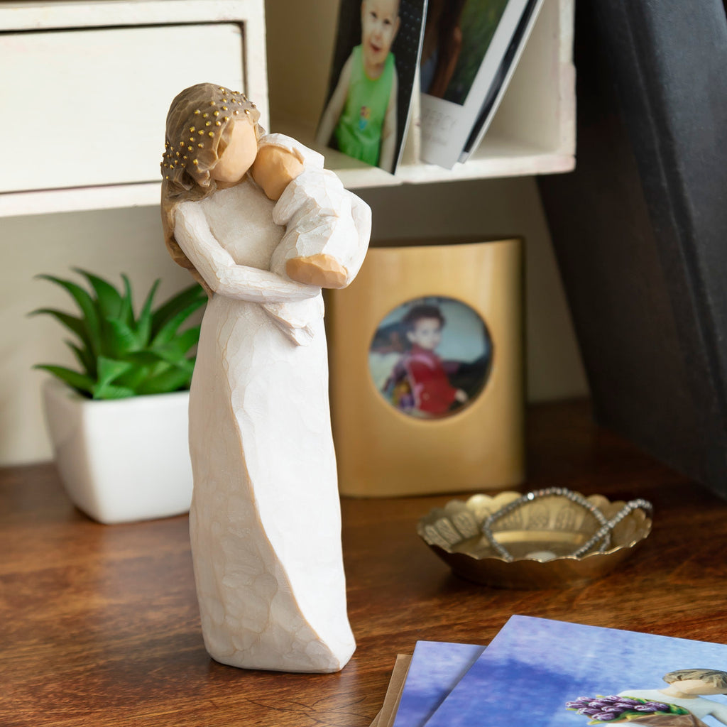 Willow Tree Sanctuary, 6.5”h hand-painted standing resin figure in cream dress, gold-leaf dots encircling her hair and face, holding infant in cream blanket, on desk with family photos