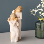 Willow Tree Loving My Mother, 6.5”h hand-painted resin figure of seated older woman with younger woman standing at her shoulder, both in cream dresses, hands clasped, on table with white flowers