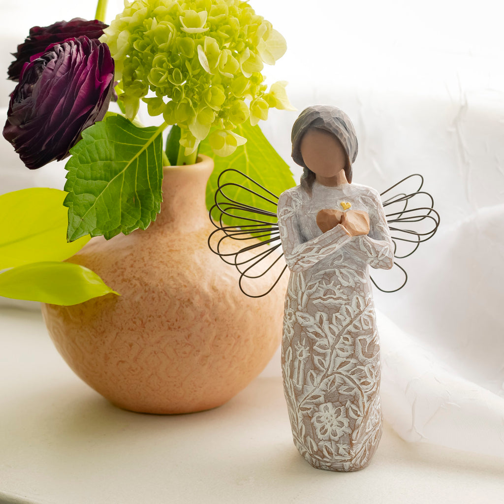 Willow Tree Remembrance (darker skin), 5.5”h hand-painted standing resin angel figure with wire wings, arms crossed over gold heart, in cream dress carved with nature icons, on table with florals