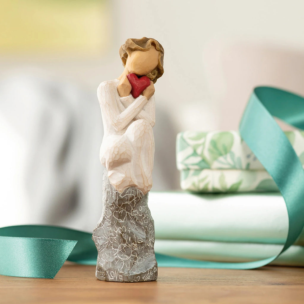 Willow Tree Always, 6”h hand-painted figure in cream dress, knees drawn up to chest, holding red heart to face, seated on gray rock etched with symbols of love, on table with teal ribbon