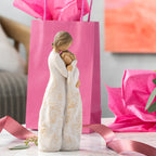 Willow Tree Close to me, 8"h hand-painted standing resin figure of woman embracing tween girl, both in cream dresses carved with intertwined vines and leaves, on table with pink gift bag