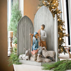 Side view of light grey barn figurine with man and woman figurine sitting in the front of it holding baby, two white sheep, one white donkey